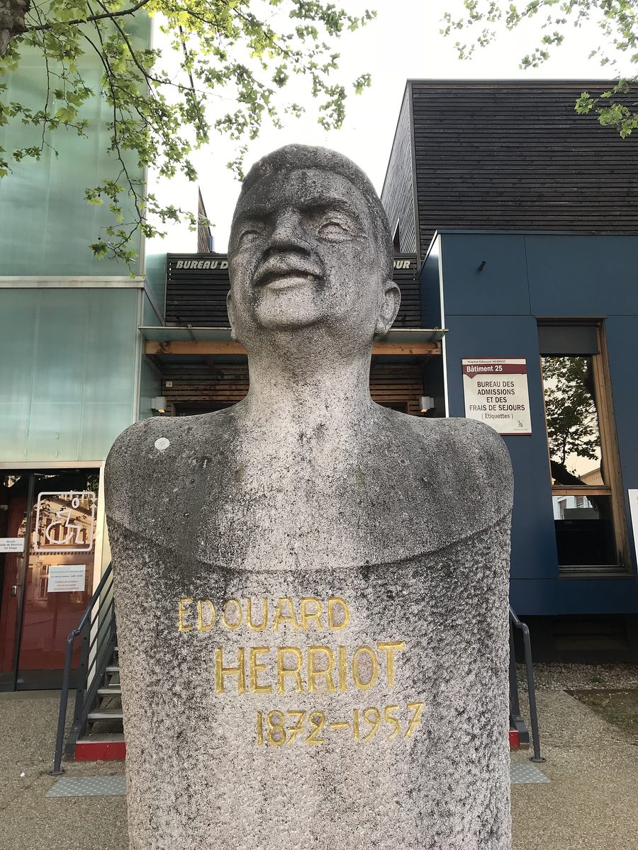 Buste engainé en pierre calcaire d’Édouard Herriot, portant l’inscription dorée Édouard Herriot 1872-1957, devant un bâtiment de l’hôpital