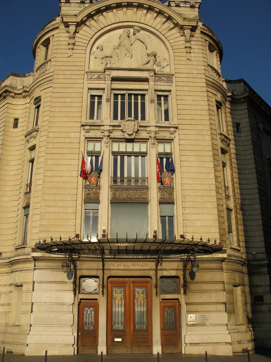 Façade en pierre du Palais de la Mutualité, avec son fronton sculpté, ses drapeaux et sa marquise au-dessus de l’entrée